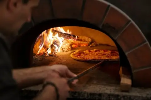 En pizzabagare använder en spade för att flytta en pizza i en vedeldad ugn. Lågorna visar den höga pizzaugn temperatur som krävs för perfekt bakning.