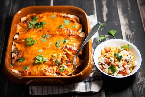 Enchiladas kyckling i en ugnsform, toppade med ost och koriander, bredvid en skål med majs- och tomatsallad.