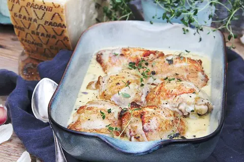 Kycklinglår i krämig sås, toppad med färska örter. En bit Grana Padano-ost syns i bakgrunden, redo att rivas över.