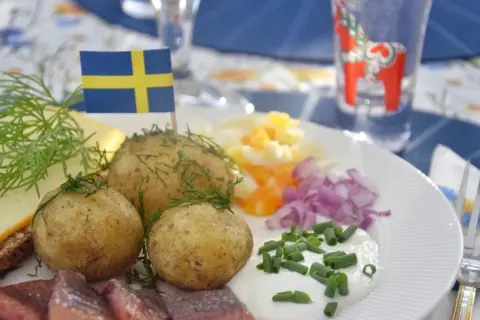 En tallrik med klassisk midsommarmat: sill, färskpotatis, ost, gräddfil och rödlök. En svensk flagga vajar stolt.