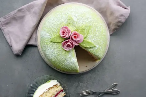 En vacker prinsesstårta med grönt marsipanlock, rosa rosor och blad. Ett stycke är uppskuret, perfekt för ett prinsesstårta recept.