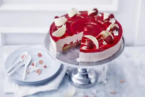 En vacker hallon cheesecake med hallonspegel, vita chokladdekorationer och färska hallon. Serveras på ett tårtfat med en skiva borttagen.