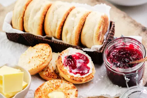 En korg med gyllene engelska muffins, en burk sylt och smör. En muffin är delad och bredd med smör och sylt.
