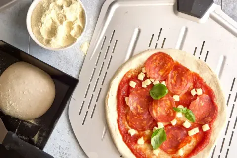 Pizzadeg till pizzasten, med pepperoni och basilika, redo att bakas. Bredvid ligger mer deg och mjöl.