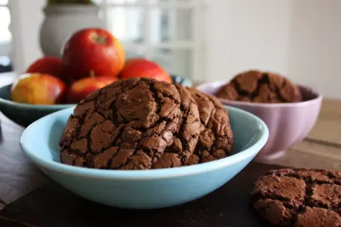 Mörka, sega kladdkake cookies i en ljusblå skål, med äpplen i bakgrunden. En annan skål med cookies syns i bakgrunden.