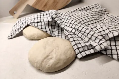 Två runda pizzadegar ligger på en bänk, en täckt av en rutig kökshandduk. Redo för bakning samma dag.