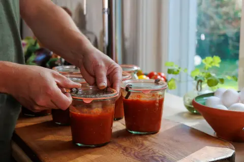 Händer som stänger glasburkar fyllda med mustig, passerad tomat. En skål med ägg och färska tomater syns i bakgrunden.
