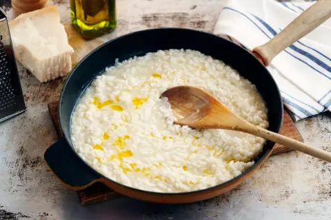 Krämig risotto i panna, med en träsked som rör om. Ingredienser som parmesan och olivolja syns i bakgrunden, redo för ett gott risotto recept.