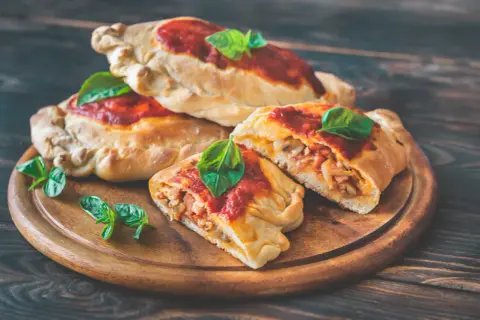 En stapel gyllene calzoner toppade med tomatsås och färsk basilika. En calzone är uppskuren och visar en fyllning av kött och grönsaker. Perfekt för den som vill veta mer om calzone kalorier.