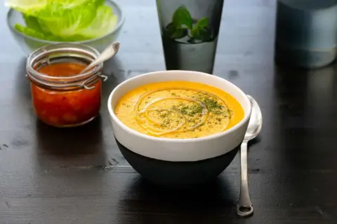En krämig linssoppa med en virvel av olja och örter. Bredvid står en burk med en röd röra och en skål med salladsblad. Ett perfekt linssoppa recept för en mysig kväll.