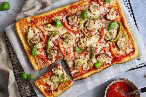 En avskuren bit av en rektangulär pizza med skinka, champinjoner och paprika. Vilken skinka till pizza är bäst? Frågan är svår att svara på, men denna ser god ut!