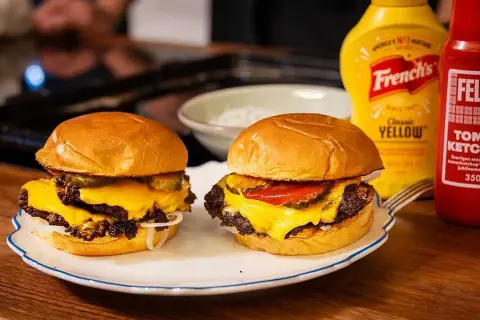 Två saftiga hamburgare med smält ost, redo att stekas. En flaska ketchup och senap står redo.