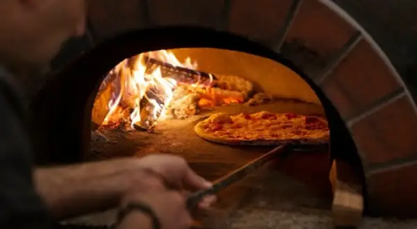 En pizzabagare använder en spade för att flytta en pizza i en vedeldad ugn. Lågorna visar den höga pizzaugn temperatur som krävs för perfekt bakning.