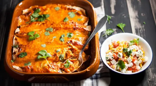 Enchiladas kyckling i en ugnsform, toppade med ost och koriander, bredvid en skål med majs- och tomatsallad.