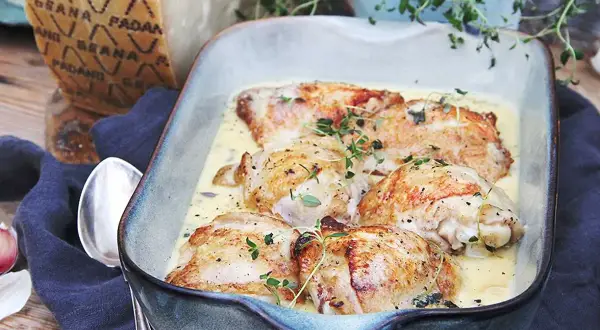 Kycklinglår i krämig sås, toppad med färska örter. En bit Grana Padano-ost syns i bakgrunden, redo att rivas över.
