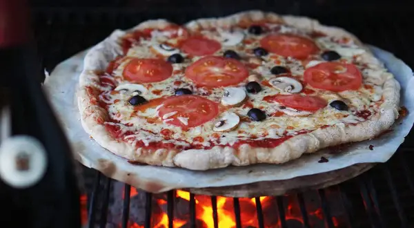 En ooni pizzadeg med tomater, champinjoner och oliver grillas över glödande kol.