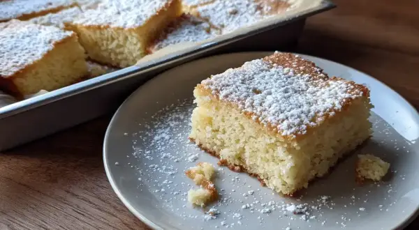 En saftig sockerkaka i långpanna, pudrad med florsocker. En bit ligger på en tallrik, redo att avnjutas.