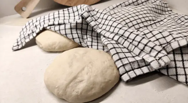 Två runda pizzadegar ligger på en bänk, en täckt av en rutig kökshandduk. Redo för bakning samma dag.