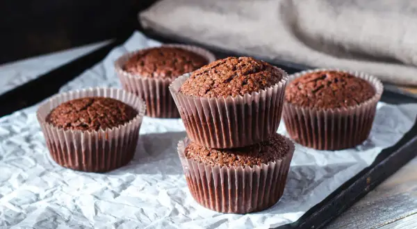 Fem choklad cupcakes på ett bakplåtspapper. Två staplade på varandra i förgrunden, med tre till utspridda runt dem.