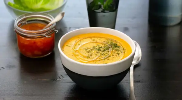 En krämig linssoppa med en virvel av olja och örter. Bredvid står en burk med en röd röra och en skål med salladsblad. Ett perfekt linssoppa recept för en mysig kväll.
