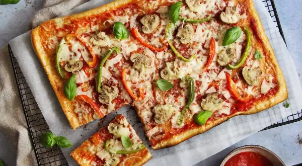 En avskuren bit av en rektangulär pizza med skinka, champinjoner och paprika. Vilken skinka till pizza är bäst? Frågan är svår att svara på, men denna ser god ut!