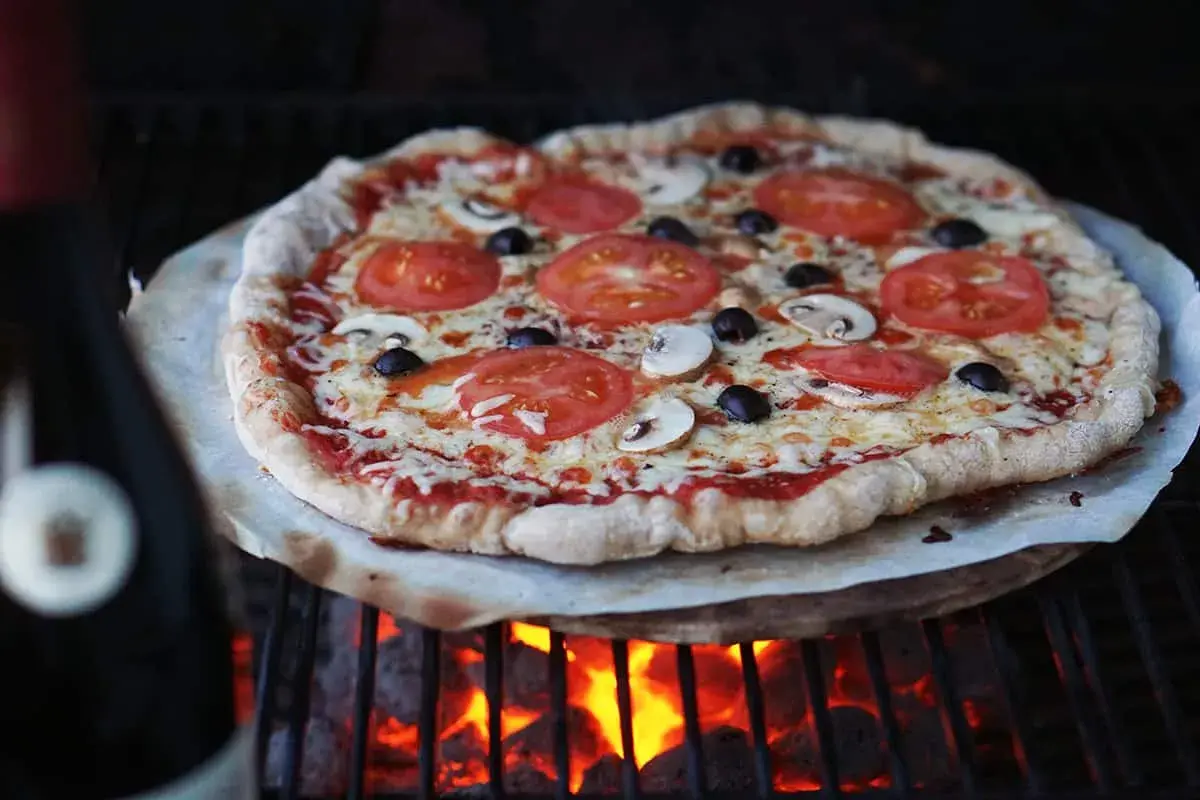 En ooni pizzadeg med tomater, champinjoner och oliver grillas över glödande kol.